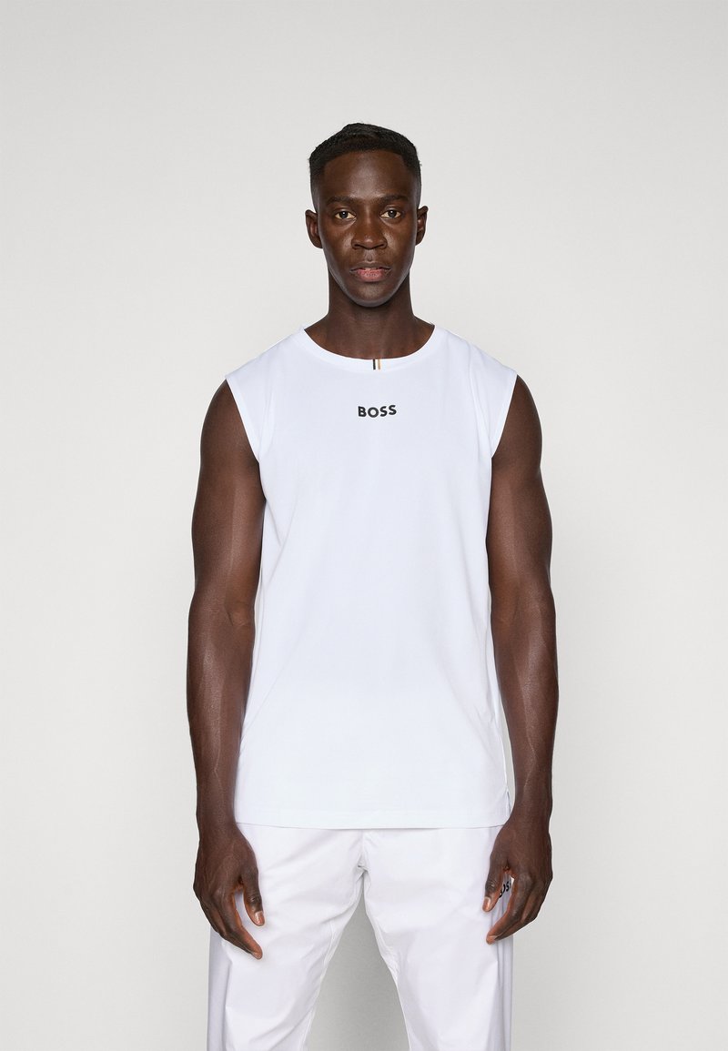 Λευκή άνετη μπλούζα χωρίς μανίκια με το λογότυπο "BOSS" σε μαύρο χρώμα, φτιαγμένη από μαλακό ύφασμα. Το μοντέλο φοράει ταιριασ�τά λευκά παντελόνια, στέκεται σε ουδέτερο φόντο.