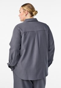 Sieviete ar blondiem matiem, kas sapīti bunā, tērpta brīvā pelēkā garām piedurknēm krekliņā un atbilstošās biksēs, stāv ar muguru pret baltu fonu.