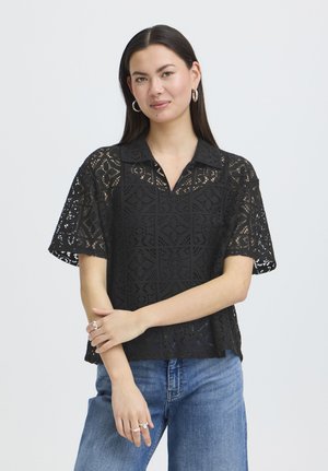 Kobieta o długich ciemnych włosach, nosząca duże koła kolczyków, czarny koronkowy krótki rękaw z kołnierzykiem oraz niebieskie dżinsy, stojąca ze skrzyżowanymi ramionami.