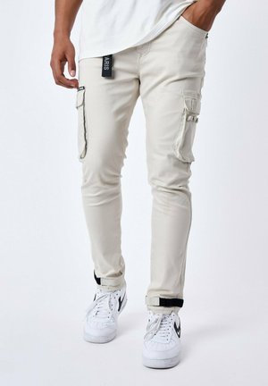 Pantalones cargo beige con bolsillos con cremallera y puños ajustables, combinados con zapatillas blancas con acentos negros. Textura de tela suave.