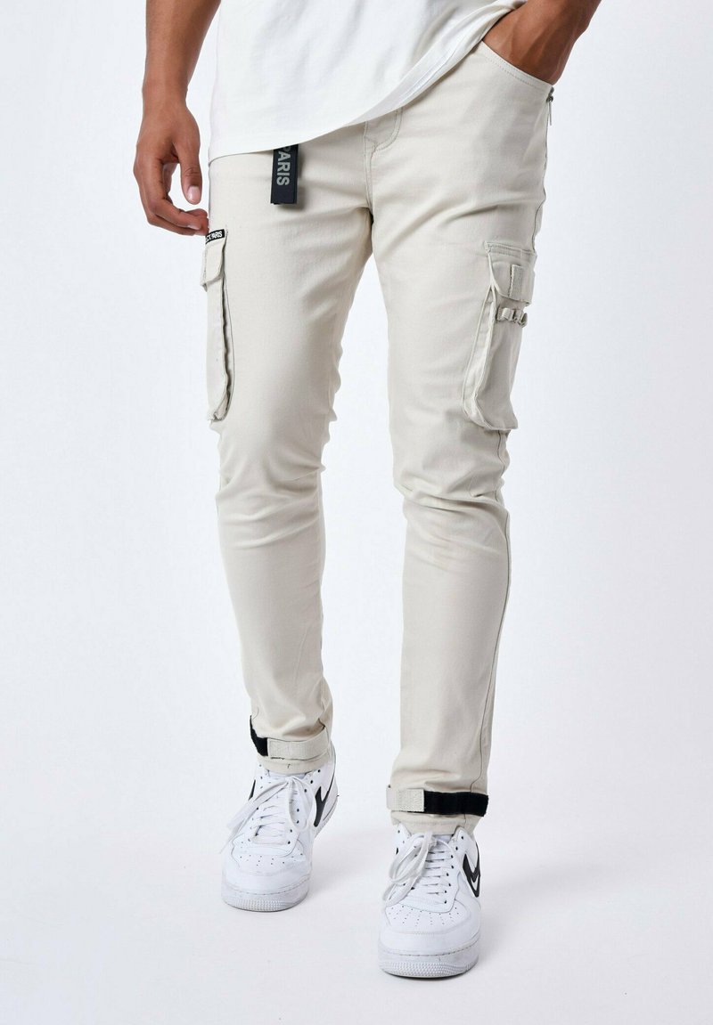 Pantalones cargo beige con bolsillos con cremallera y puños ajustables, combinados con zapatillas blancas con acentos negros. Textura de tela suave.