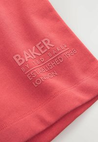 Koraalstof met een gladde textuur heeft reliëftekst: "BAKER by TED BAKER OPGERICHT 1988 LONDEN" in lichtroze.