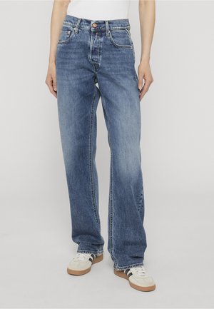 Personne portant un jean bleu coupe décontractée et des baskets blanches avec des bandes noires, debout devant un fond clair uni.
