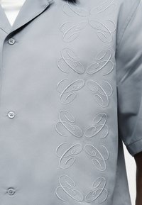 Ανοιχτό γκρι κοντομάνικο πουκάμισο με κάθετο κεντητό σχέδιο στροβίλου στη δεξιά πλευρά, φορεμένο πάνω από λευκό τοπ.