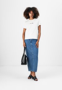T-shirt en coton blanc avec broderie "Sunny Happy", associé à une longue jupe en denim, des sandales à talons blocs noires et un sac à main noir.
