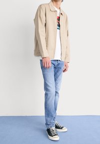 Hombre con chaqueta beige sobre camiseta blanca con estampado, jeans azul claro y zapatillas negras, de pie sobre un suelo azul frente a una pared blanca.