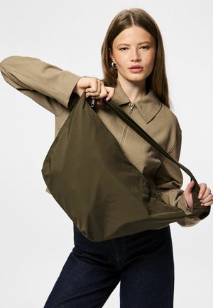 Oliwkowa nylonowa torba o gładkiej fakturze, wyposażona w długi pasek i otwarty górny design. Model trzyma torbę, podkreślając jej rozmiar.