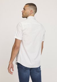Chemise à manches courtes, blanche avec des motifs subtils géométriques, mettant en avant un tissu doux. Présente un col standard et un ourlet arrondi.
