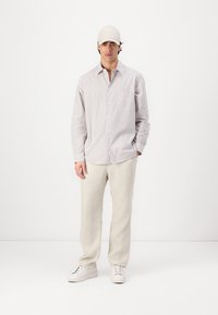 Chemise à rayures dans des couleurs claires, pantalon beige clair et baskets blanches, assortis d'une casquette beige, sur un fond uni.