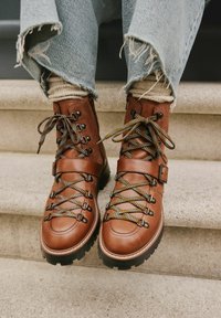 Braune Lederwanderschuhe mit schwarzen Gummisohlen, ausgestattet mit mehrfarbigen Schnürsenkeln und einem Knöchelriemen, getragen mit ausgefransten Jeans.