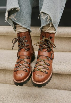 Brown leather hiking boots with black rubber soles, featuring multi-colored laces and an ankle strap, worn with frayed denim jeans.
