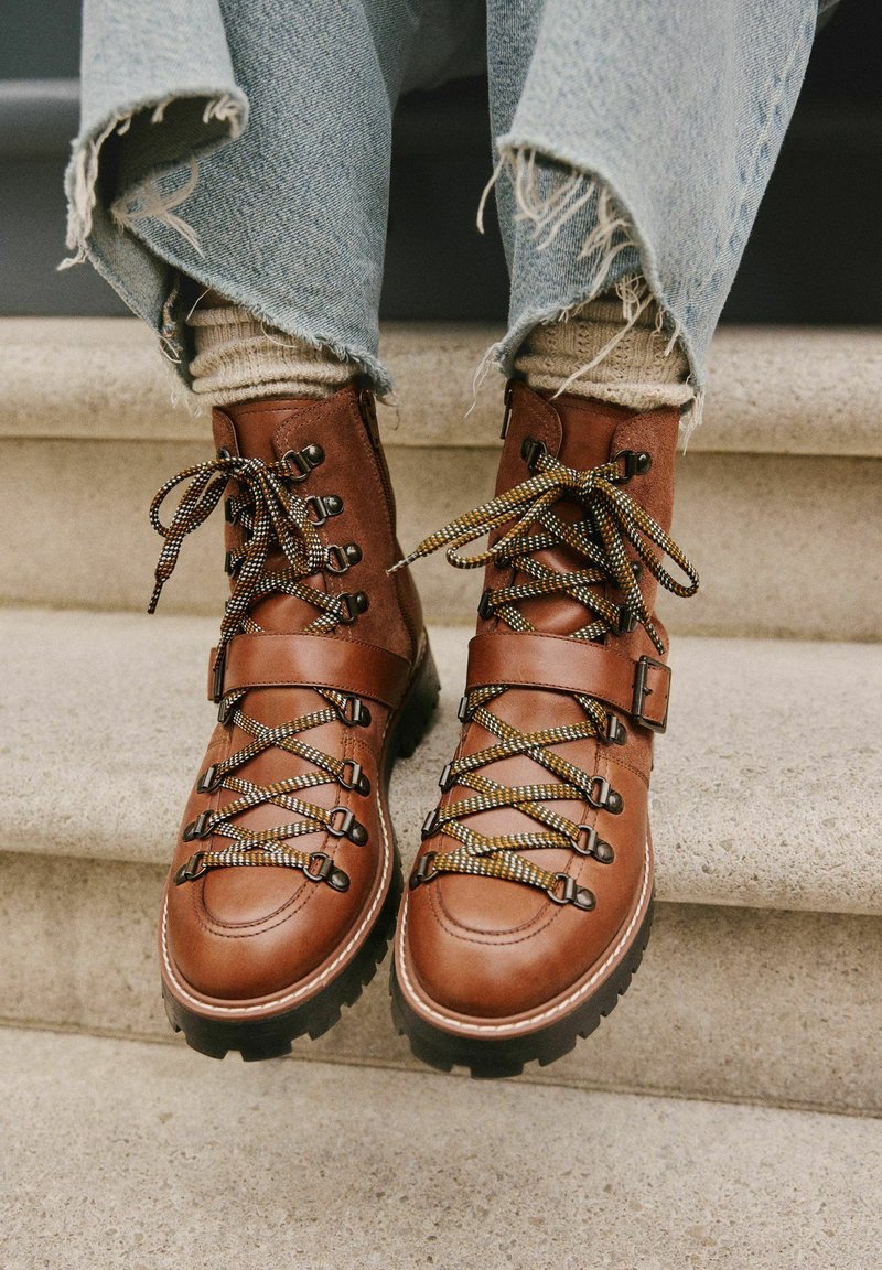 Braune Lederwanderschuhe mit schwarzen Gummisohlen, ausgestattet mit mehrfarbigen Schnürsenkeln und einem Knöchelriemen, getragen mit ausgefransten Jeans.