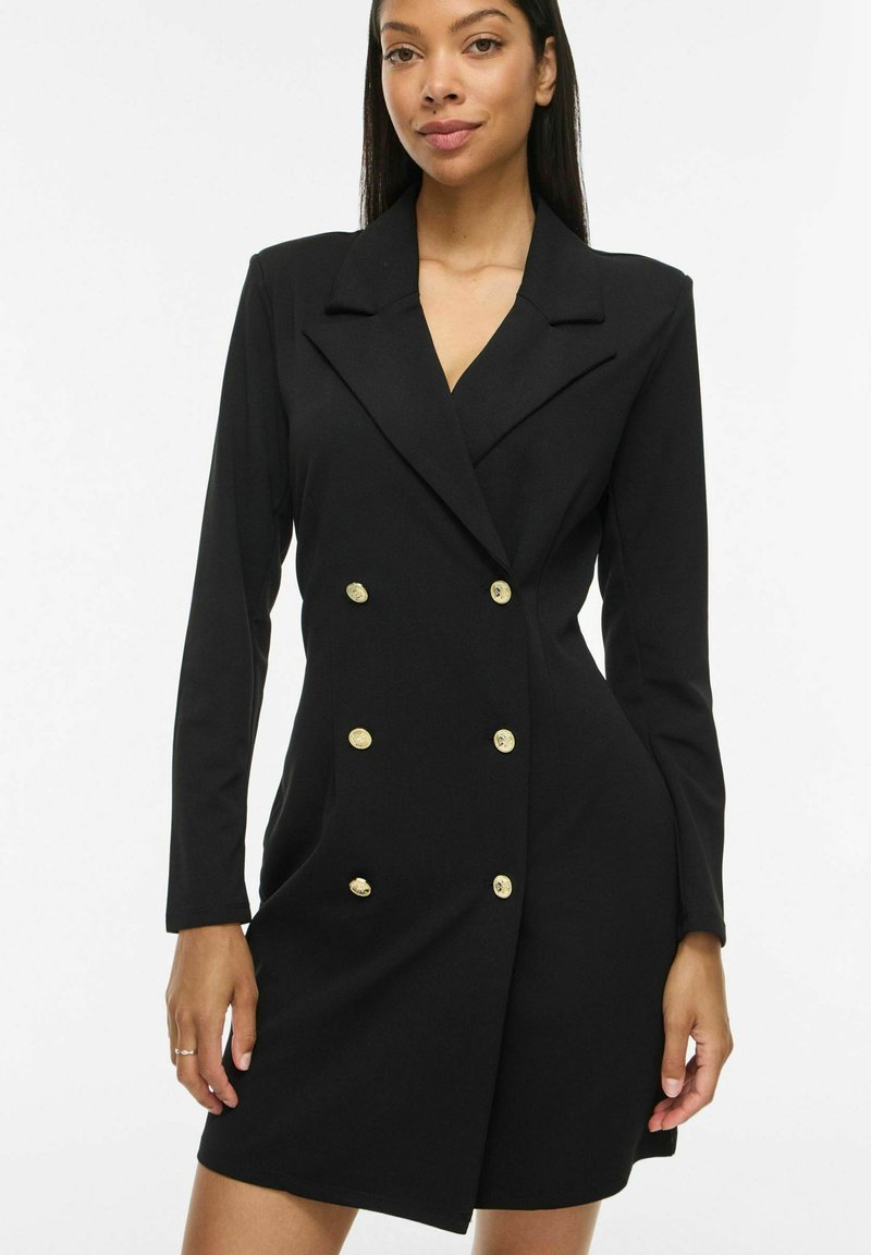 Femme portant une robe blazer noire cintrée avec des boutons dorés, manches longues et col cranté, debout devant un fond blanc uni.