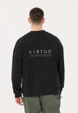 Mann, der von hinten zu sehen ist, trägt einen schwarzen Pullover mit großem "VIRTUS"-Schriftzug und kleinem beschreibendem Text auf dem Rücken, kombiniert mit grünen Hosen.