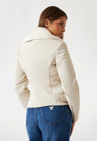 Jachetă din piele albă cu guler din blană sintetică, detalii matlasate pe laterale și talie cu curea. Purtați-o cu blugi din denim albastru.
