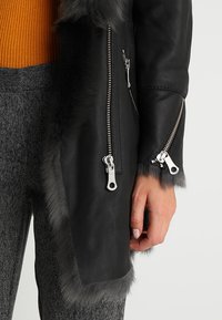 Sort læderjakke med grå pelskant, der har sølvlynlåse og en tætsiddende silhuet. Struktureret ribbet orange sweater under.