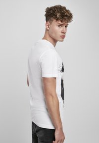 White cotton t-shirt with short sleeves, featuring a black graphic print on the front. Model poses sideways, wearing dark jeans.