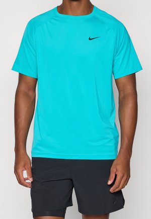 Mężczyzna ubrany w turkusową sportową koszulkę z krótkim rękawem z czarnym logo Nike i czarne sportowe spodenki na jednolitym tle.