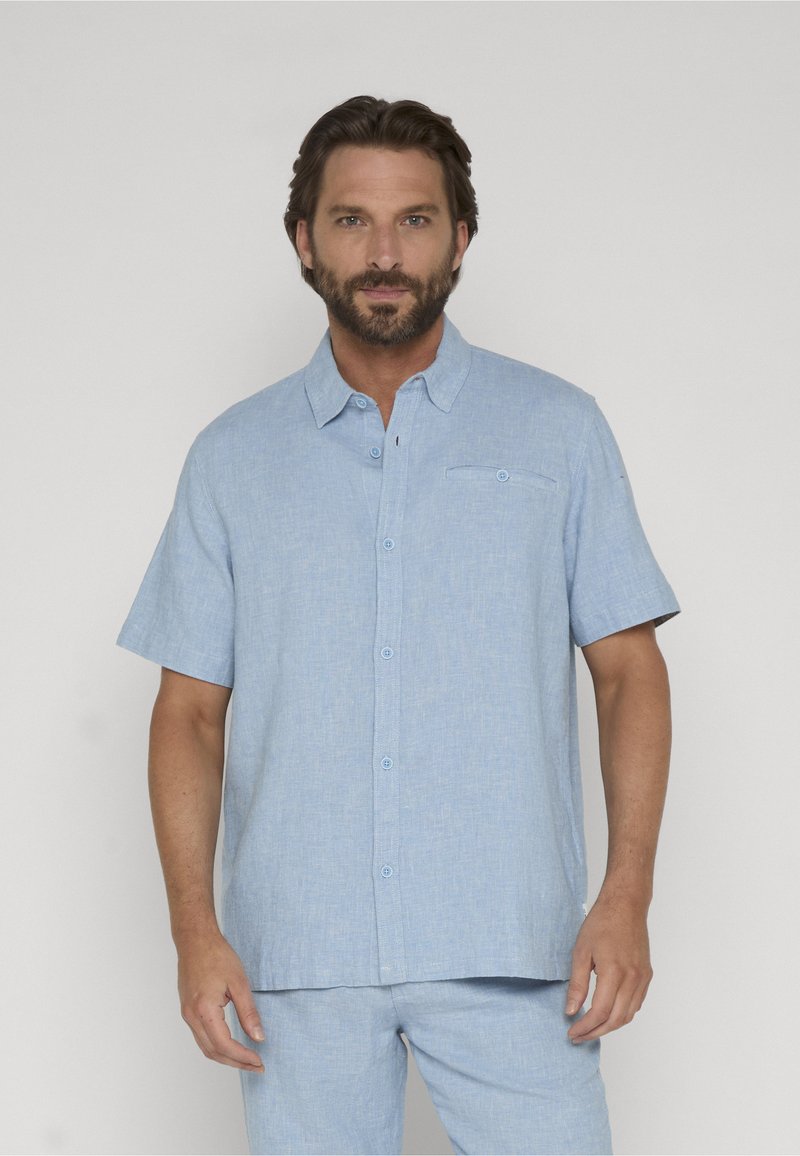 Homme barbu portant une chemise à manches courtes bleu clair boutonnée et un pantalon assorti sur un fond uni.