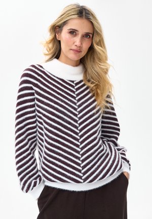 Blondynka z luźnymi lokami, ubrana w czarno-biały pasiasty sweter z białym kołnierzykiem, ręce w kieszeniach ciemnych spodni, neutralne tło.