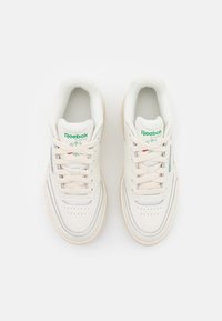 Pár bílých tenisek Reebok Classic se zeleným logem, béžovými tkaničkami a perforovanými detaily, zobrazených shora na bílém pozadí.