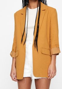 Personne portant un blazer moutarde avec des manches retroussées, un t-shirt blanc à côtes et un short blanc à l'ourlet effiloché, avec de longues tresses noires visibles.