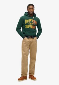 Zielona bawełniana bluza z kapturem z grafiką gór i napisem "Superdry", zestawiona z beżowymi spodniami i brązowymi sk�órzanymi butami. Bluza ma sznureczki w kapturze.