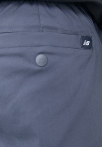 Jūras zila auduma šorti ar pogu aizdari un aizmugurējo kabatu, kurā ir neliela New Balance logo etiķete. Gluda, teksturēta auduma virsma.