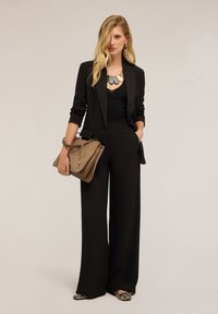 Completo nero su misura con pantaloni a gamba larga e una parte superiore aderente. Gli accessori includono una collana a più strati e una borsa color nocciola. Scarpe in stampa serpente.