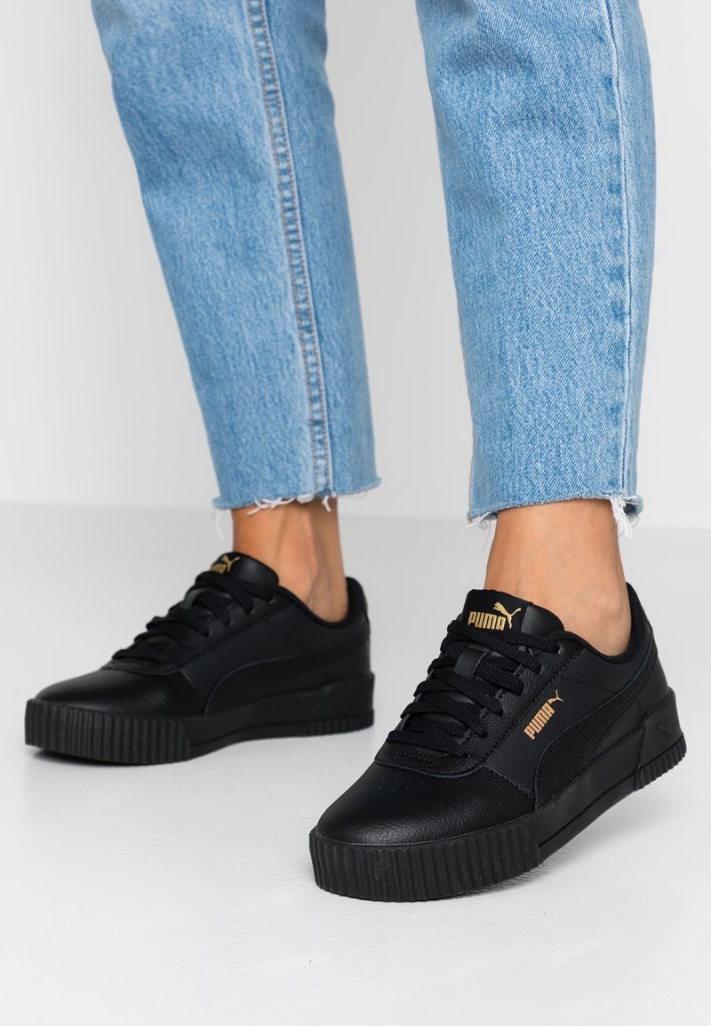 Baskets noires Puma avec logo doré portées avec un jean bleu clair coupé au-dessus de la cheville sur un sol blanc.
