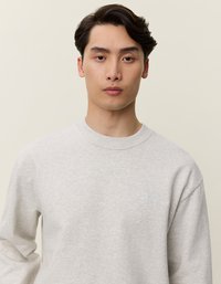 Pilkas megztinis su apvaliu kaklu, raukšlėtais rankogaliais ir mažuoju baltu logotipu kairėje krūtinės pusėje. Minkšta medžiaga su lygiu paviršiumi.