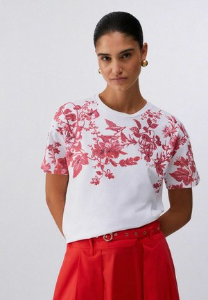 Donna con capelli lisciati all'indietro, indossa una maglietta bianca con stampa floreale rossa e pantaloni rossi a vita alta con cintura, su uno sfondo chiaro.