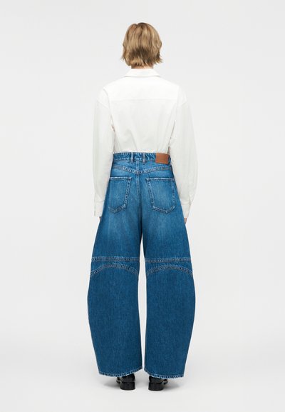 Άτομο με κοντά ξανθά μαλλιά φοράει λευκό πουκάμισο με μακριά μανίκια, μπαίνει μέσα σε ψηλόμεσα φαρδιά μπλε τζιν, στεκόμενο μπροστά σε απλό φόντο.