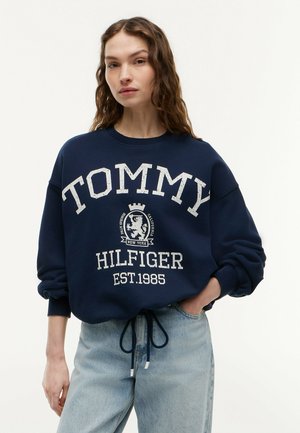 Kobieta w granatowej bluzie Tommy Hilfiger i jasnoniebieskich jeansach, stojąca z jedną ręką w kieszeni na jednolitym tle.