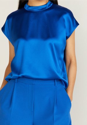 Frau trägt eine kobaltblaue satinierte Kurzarmbluse, die in eine passende, maßgeschneiderte Hose mit hoher Taille gesteckt ist, die Hände in den Taschen.