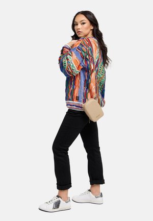 Vrouw in een kleurrijke trui met patroon, zwarte opgerolde broek, witte sneakers en een kleine beige crossbodytas, poseert zijwaarts tegen een witte achtergrond.