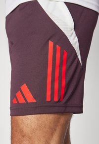 Pantalones cortos atléticos morados con acentos contrastantes en blanco y rojo. Presenta tres franjas diagonales rojas y un diseño geométrico rojo en el lateral.