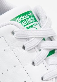 White Adidas Stan Smith sneaker with white laces tied over a green and white branded tongue label.