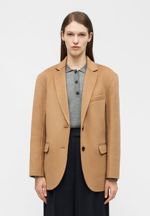 Žena s rovnými hnedými vlasmi, oblečená v kamelovom saku cez sivú košeľu na gombíky a čierne nohavice, na pozadí jednoduchého bieleho pozadia.