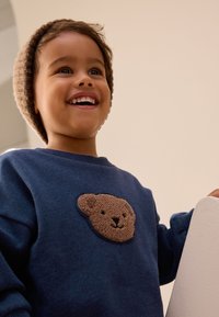 Tengerészkék pulóver barna maci mintával a mellkason. Gyerek világosbarna kötött sapkában, mosolyog.