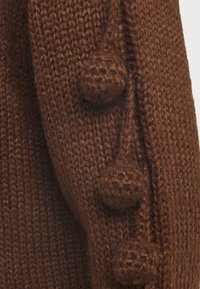 Section d'un pull en tricot marron avec un fil texturé, agrémentée de trois pompons ronds et surélevés sur le côté.
