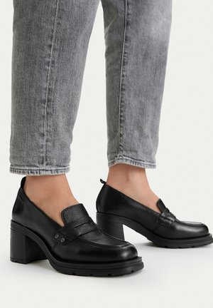 Person trägt graue Jeans und schwarze, klobige Loafers mit Lederabsatz und steht vor hellem Hintergrund.