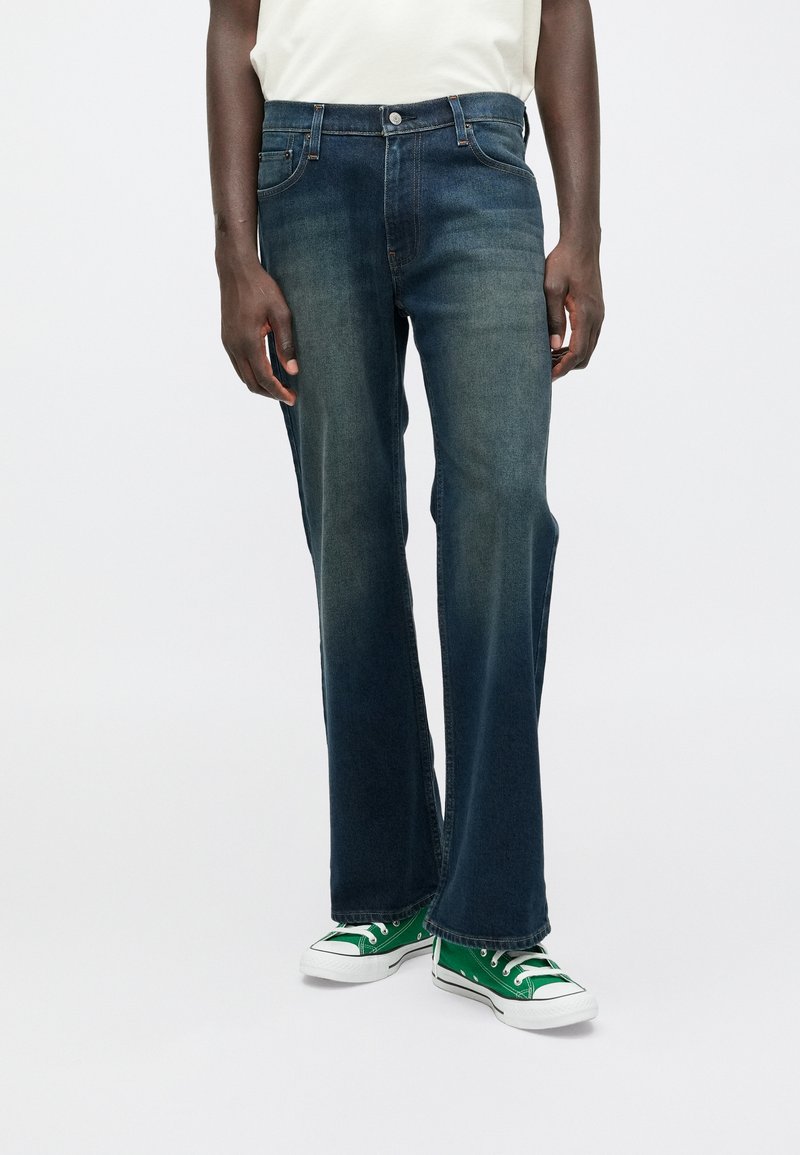 Σκούρα μπλε, ελαφρώς ξεθωριασμένα τζιν με κανονική γραμμή, σχέδιο πέντε τσεπών και ασημένιο κουμπί. Συνδυασμένα με πράσινα Converse αθλητικά παπούτσια.