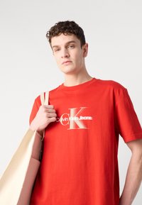 Crvena pamučna majica s bijelim i zlatnim logotipom "Calvin Klein Jeans". Model nosi bež torbu s strukturiranim dizajnom.