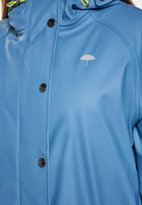 Mėlyna atspari vandeniui striukė, turinti lygų paviršių, juodus mygtukus ir subtilų pilką skėčio logotipą ant krūtinės. Reguliuojama kapušoną su žaliais akcentais.