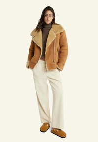 Bruine schapenvachtjas met een brede kraag, ritssluiting en lange mouwen. Gecombineerd met crèmekleurige corduroypantalons en bruine instappers.