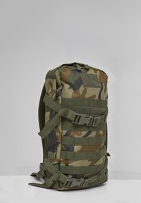 Taktinė kuprinė su maskuotės raštu, keliais dirželiais, sagtimis ir MOLLE dirbiniu daiktams pritvirtinti, stovinti vertikaliai ant lygaus paviršiaus.