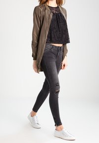 Bomber jacket verde oliva, top corto de encaje negro, jeans ajustados negros desgastados y zapatillas blancas. La chaqueta tiene puños y dobladillo acanalados.
