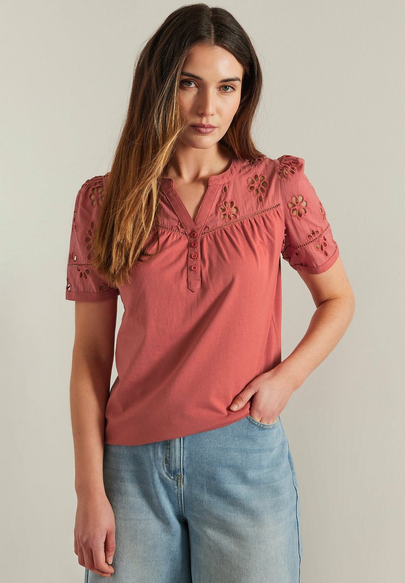 Top color rosa con dettagli floreali ricamati, maniche corte a sbuffo, scollatura a bottoni tipo henley e vestibilità rilassata. Indossato con jeans blu chiaro.