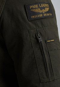 Veste vert olive en tissu texturé, avec un écusson portant la mention "PME LEGEND" et "FREEDOM UNLIMITED," et une poche zippée sur la manche.